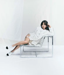 Millie Brady wearing a white textured jacket and white shoes, reclining on a modern chrome frame chair against a white seamless backdrop.