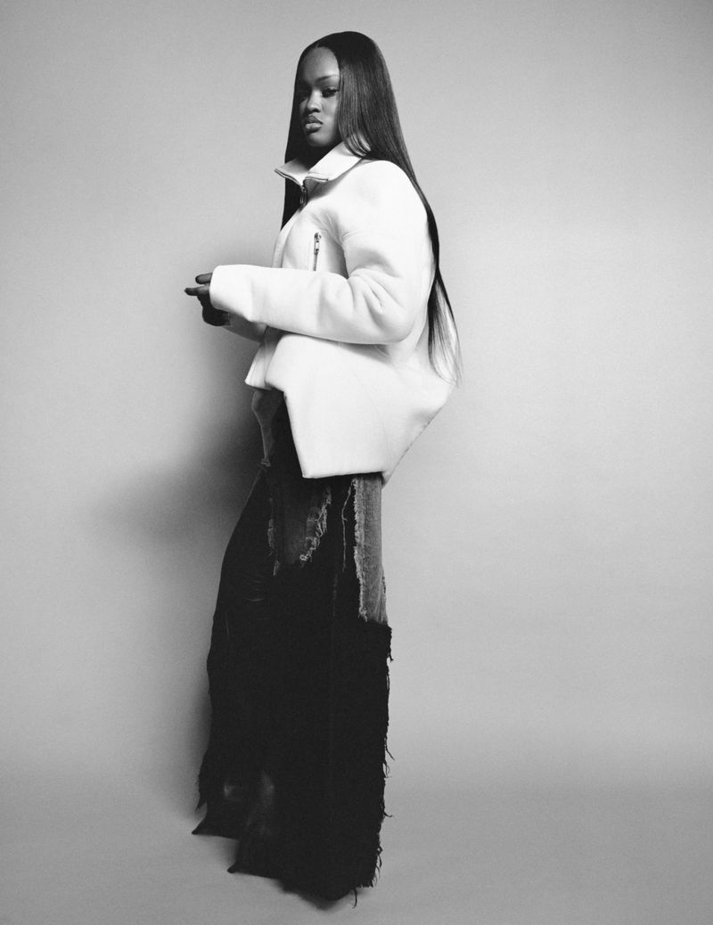 A woman with long dark hair poses in a white jacket and black skirt in a studio, captured in a black and white, medium shot.