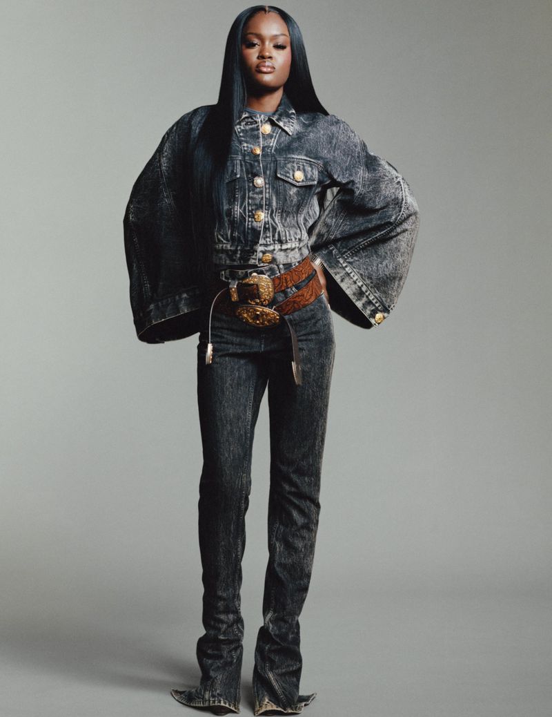 A full shot of an African American woman in an acid-wash denim outfit with a wide belt, posing in a photo studio with a neutral background.