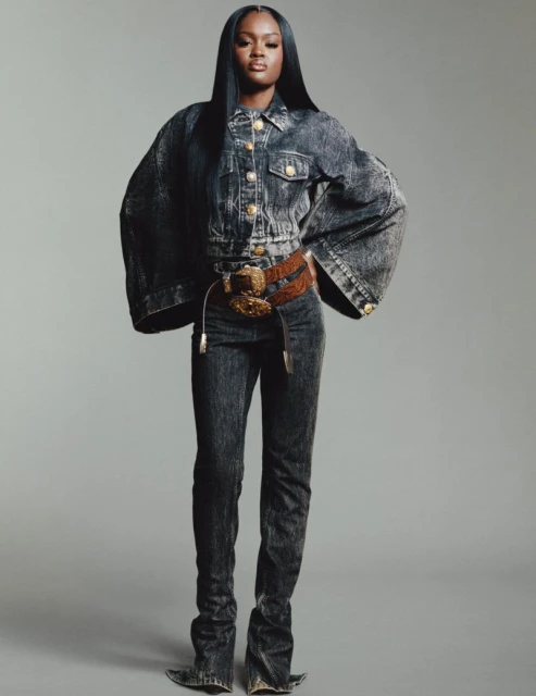 A full shot of an African American woman in an acid-wash denim outfit with a wide belt, posing in a photo studio with a neutral background.