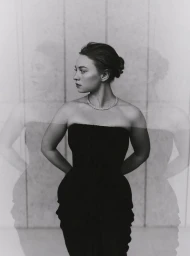 Black and white portrait of Mia Threapleton wearing a strapless dark dress and necklace against a paneled wall with multiple exposure ghosts