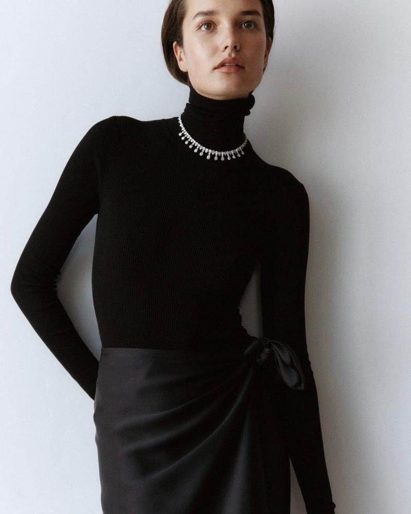 Elegant woman poses in black turtleneck sweater and black wrap skirt, adorned with a delicate diamond necklace against a neutral wall.