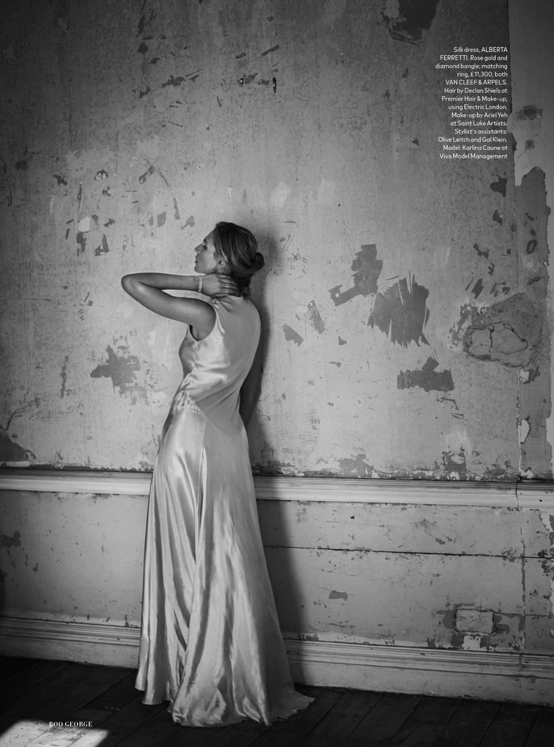 Woman in floor-length gown, with her hand at her neck, stands in a monochromatic fashion shot against a distressed wall backdrop.