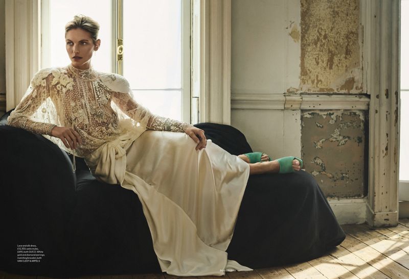 Fashion shot of a blond woman in a lace top and satin skirt reclining on a black sofa near a sunlit window in a distressed room.