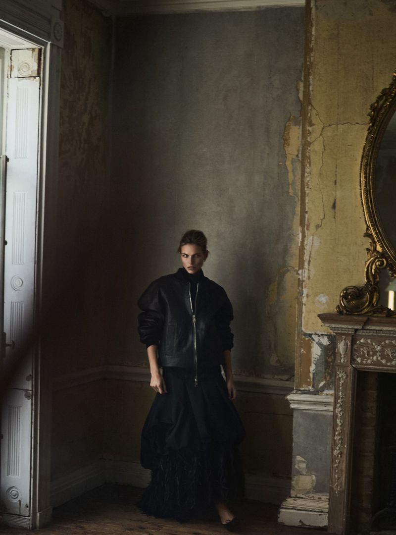 Woman in a black jacket and skirt poses in an ornate interior with distressed walls and a large mirror, creating an editorial fashion image.