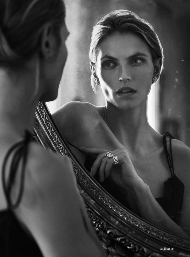 Black and white portrait of an adult female looking at her reflection in an ornate mirror; she is wearing a ring and earrings.