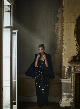 Indoor fashion shot of a woman in a black strapless dress with silver dots, black blazer, posing in an empty room with light and shadows.