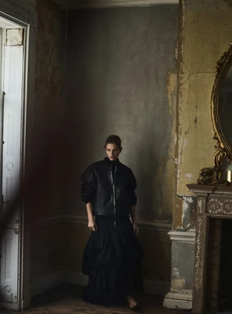 Model wearing a black jacket and long black skirt standing in an interior room with a weathered wall and an ornate fireplace.