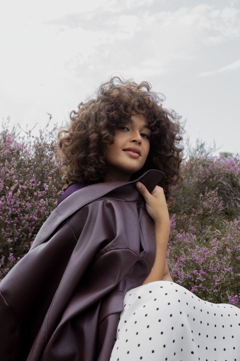 Young woman with curly hair, wearing a burgundy jacket and polka dot skirt, poses outdoors among violet flowers against a cloudy sky.