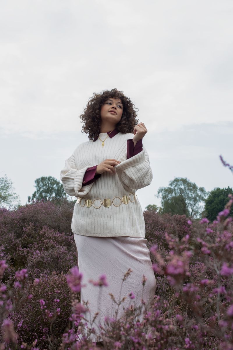 Woman with curly hair in a cream sweater and gold belt stands in a field of purple flowers under an overcast sky.
