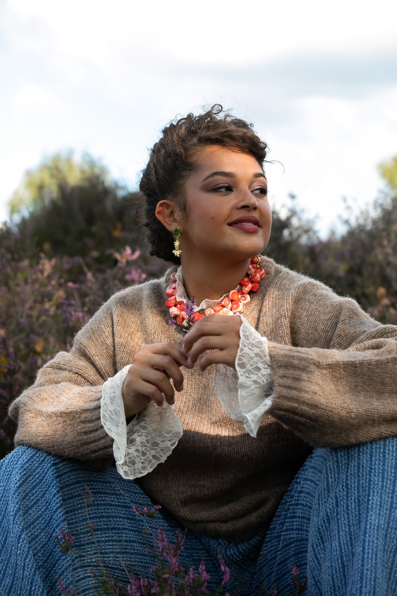 Woman in sweater and blue skirt sits in a field of lavender, wearing a colorful necklace and earrings, looking to the side.