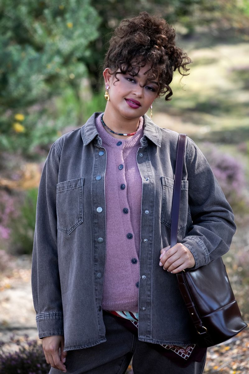 Young woman with curly hair poses outdoors in a grey shirt jacket, pink cardigan, and brown bag, set against a blurred natural background.