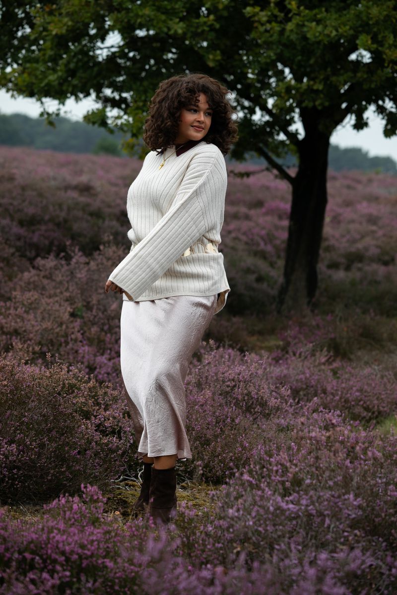 Adult female in outdoor setting wearing cream sweater, gold belt, and brown boots standing in a field of purple flowers next to a tree.