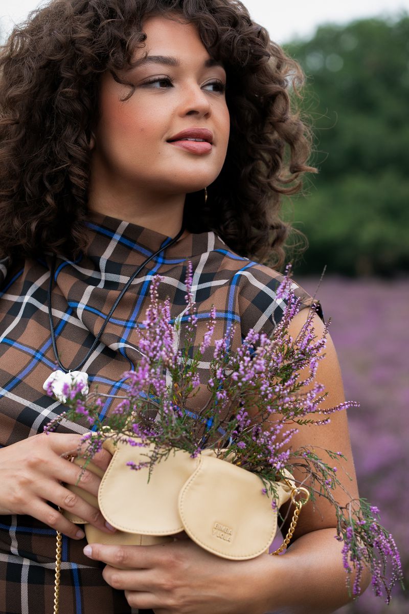 Brunette woman in a plaid top holds a small beige purse with lavender flowers, set against a blurred backdrop of heather.