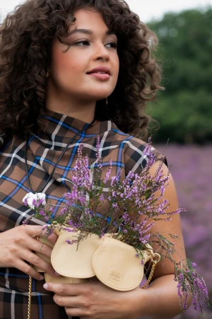 Brunette woman in a plaid top holds a small beige purse with lavender flowers, set against a blurred backdrop of heather.