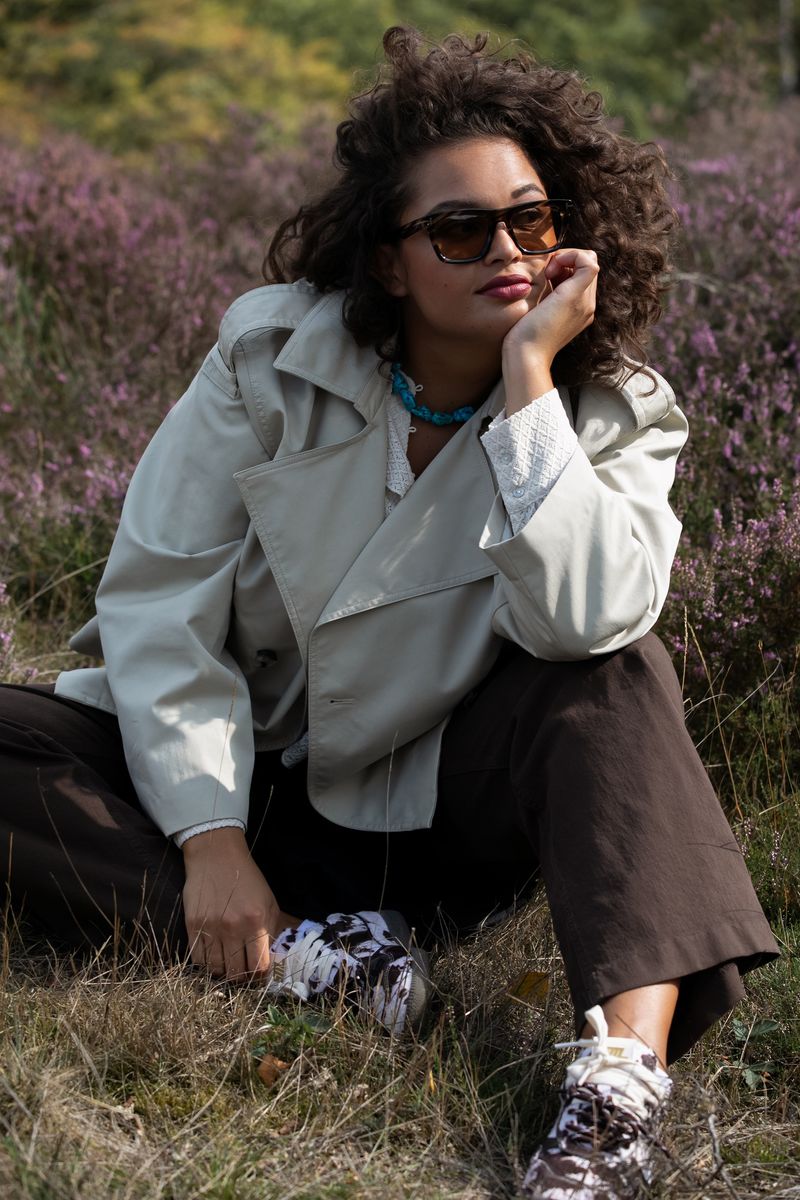 Young woman with curly hair sits in a field of purple wildflowers wearing a beige trench coat, sunglasses, and colorful sneakers outdoors.