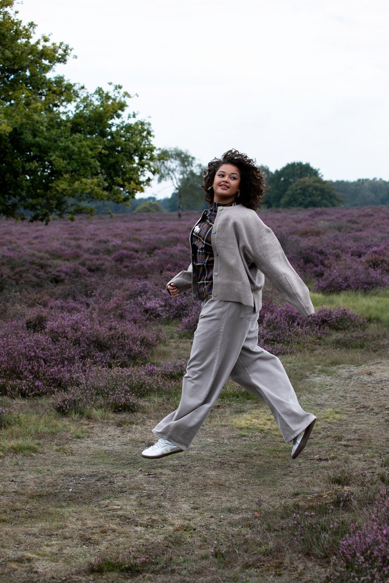 A woman with curly hair jumps in a field of heather wearing gray pants, a gray cardigan, and white sneakers in an outdoor setting.
