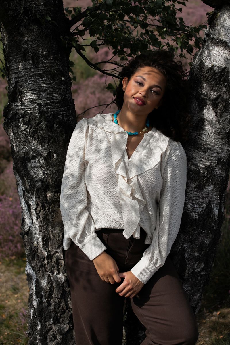 Adult female posing in an outdoor setting, wearing a white patterned ruffle shirt and brown trousers, leaning against a birch tree.