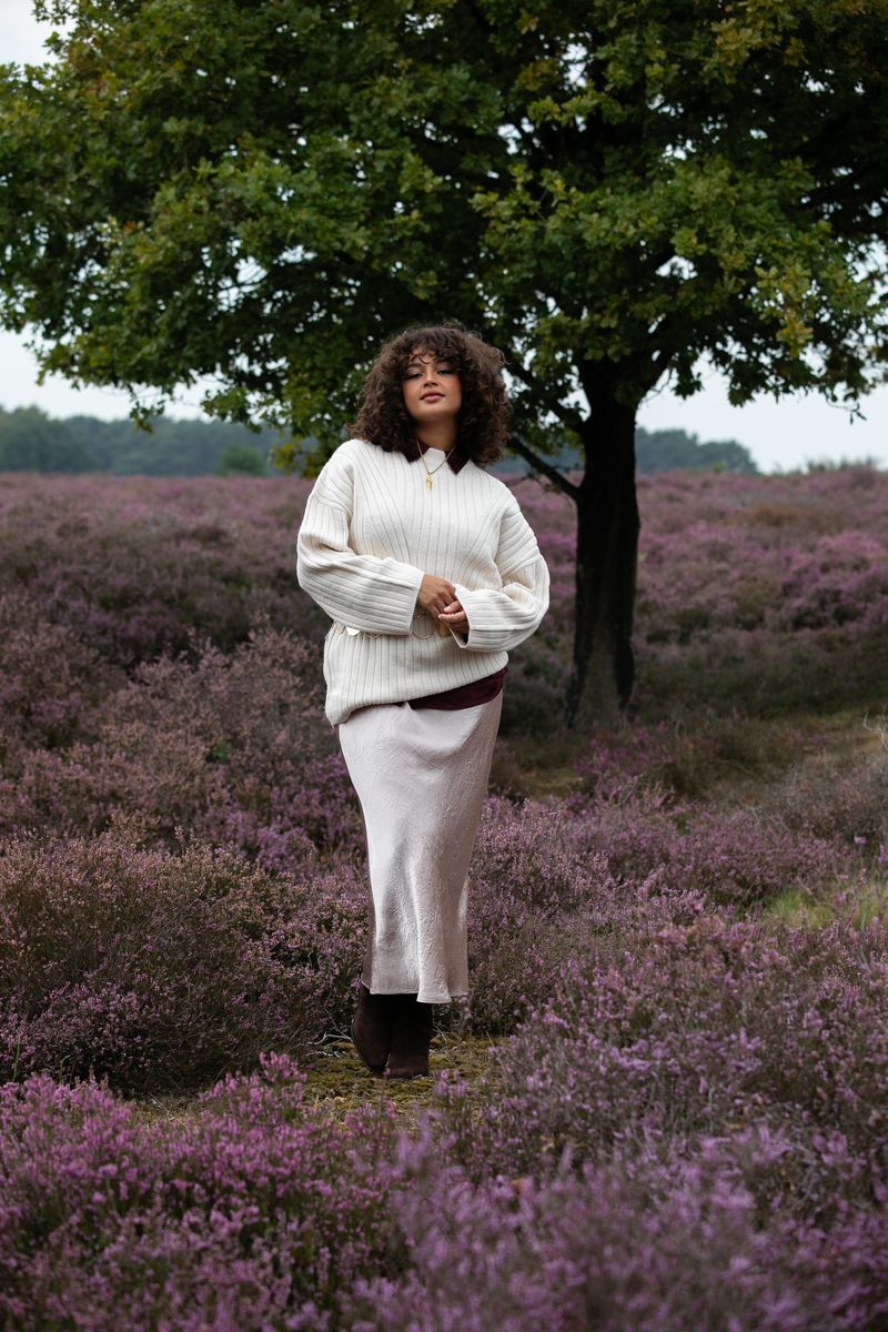 Woman with curly hair in a cream sweater and skirt standing in a field of heather with a green tree in the background.