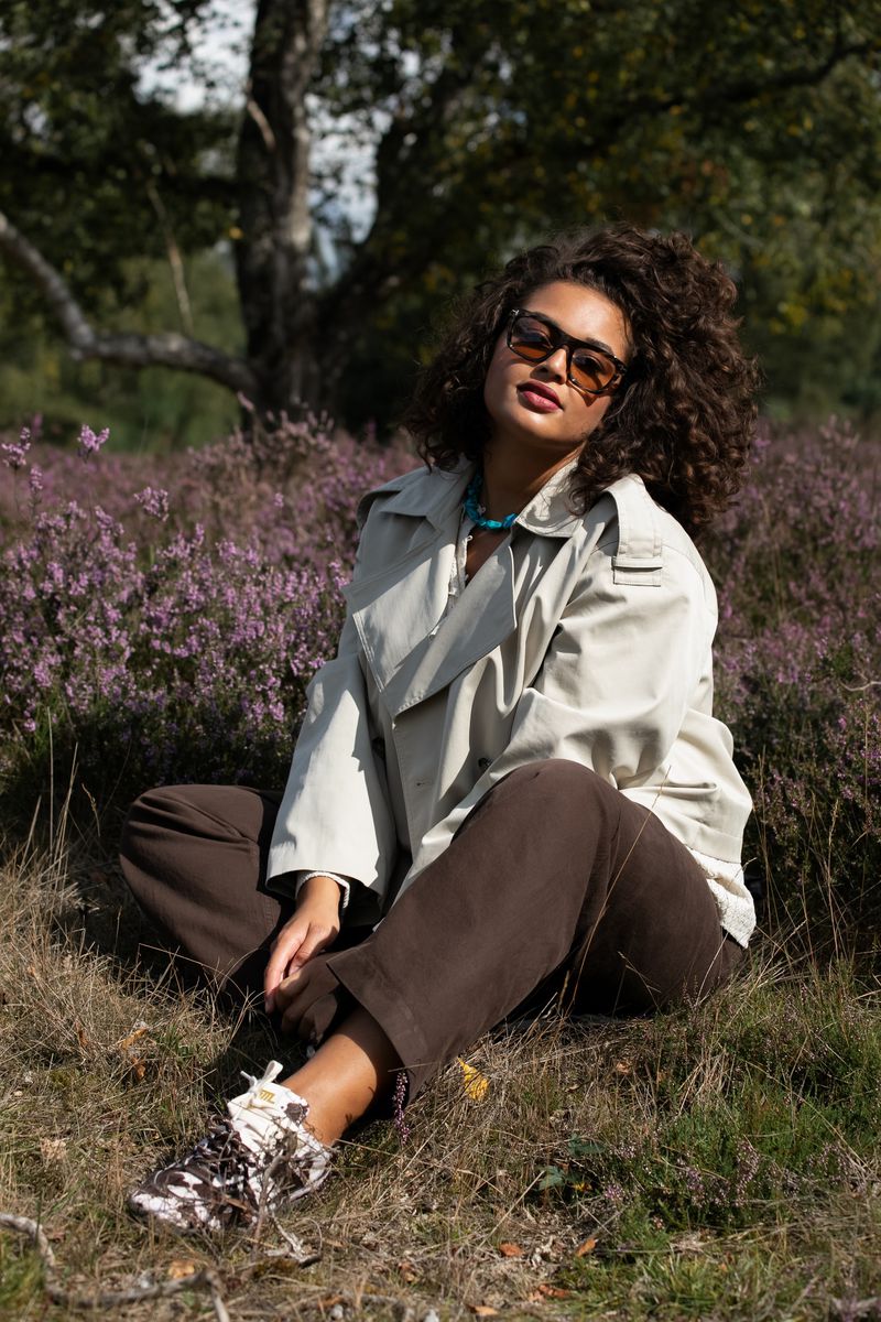 A young woman with curly hair sits cross-legged in a field of purple flowers, wearing a light trench coat and brown pants, smiling slightly.