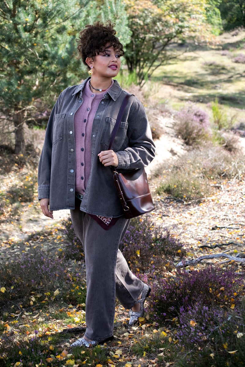 Woman in grey corduroy outfit walking in sunlit forest. She carries a shoulder bag and wears silver shoes, with trees and foliage in background.