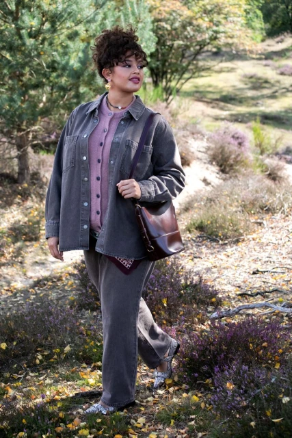Woman in grey corduroy outfit walking in sunlit forest. She carries a shoulder bag and wears silver shoes, with trees and foliage in background.