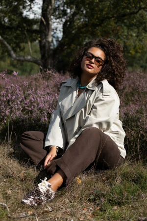 Woman with curly hair wearing sunglasses, a beige jacket, and brown trousers seated outdoors in a field of purple heather.