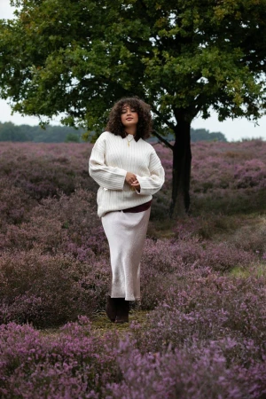 Woman with curly dark hair wearing a cream knit sweater and long skirt standing in a field of purple heather.