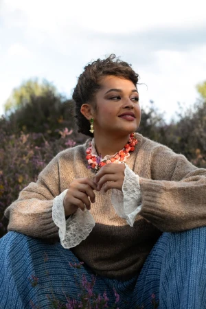 Young woman in a beige knit sweater and lace cuffs seated outdoors in a field of flowering purple heather.