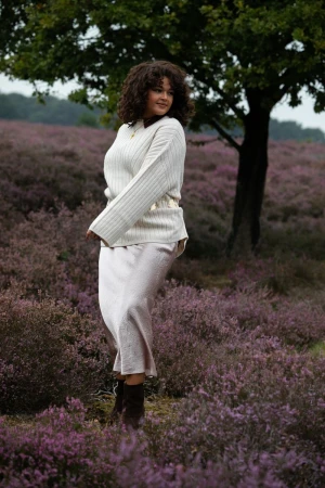 Woman with curly dark hair wearing a ribbed white sweater and skirt standing amidst purple heather flowers