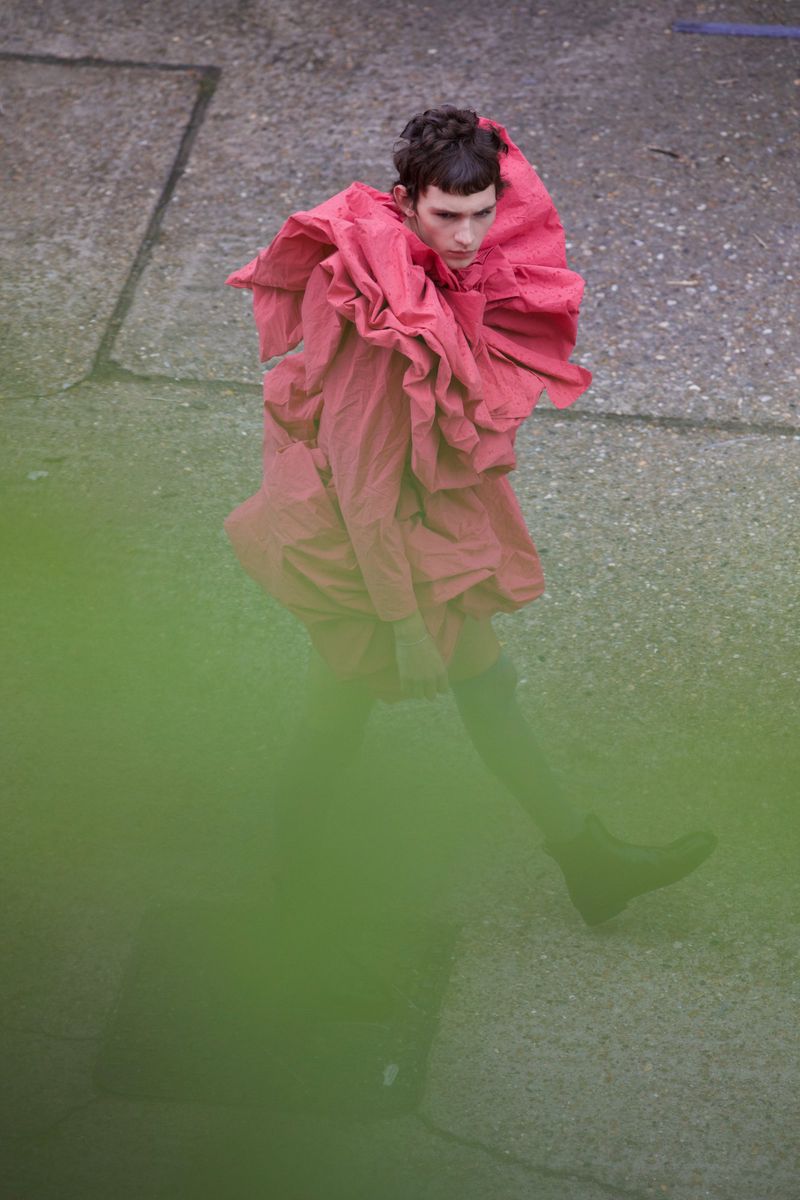A young adult in an avant-garde pink ruffle coat poses on concrete in an eye-level shot with blurred green foreground elements.
