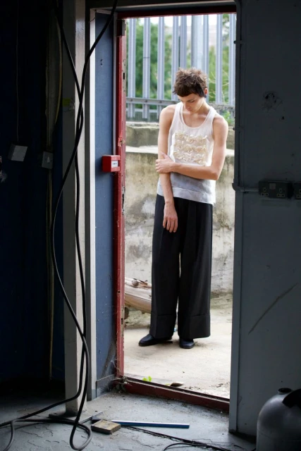 An adult female stands in the doorway of an abandoned building, wearing a tank top and slacks in a medium shot on a cloudy day.