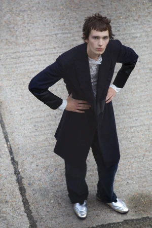 Young man in an oversized dark suit and silver shoes stands in an urban setting, posing with hands on his hips, captured from a high angle.