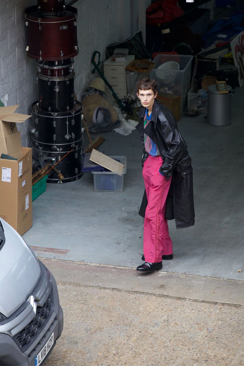Person in black coat and pink pants stands in cluttered garage near gray van and stacked drums, creating an edgy fashion scene.