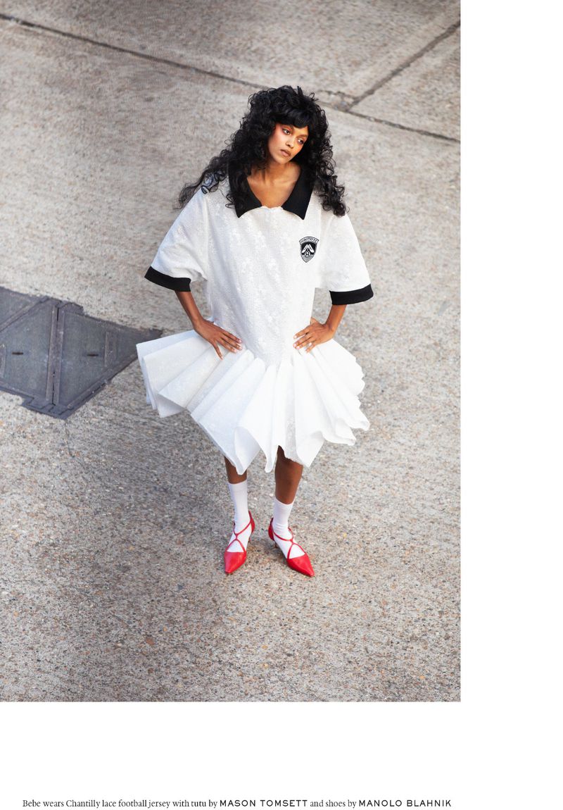 Fashion editorial portrait of a woman in a white jersey dress and red heels, posing in an outdoor urban setting on a sunny day.