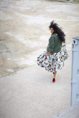 Woman with curly hair in an olive jacket and patterned skirt looks over her shoulder outdoors in a fashion style shot. 