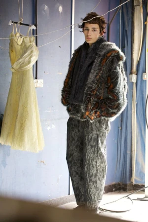 A person in a fur coat and patterned pants stands indoors next to a hanging dress. Moody lighting emphasizes textures and unique fashion.