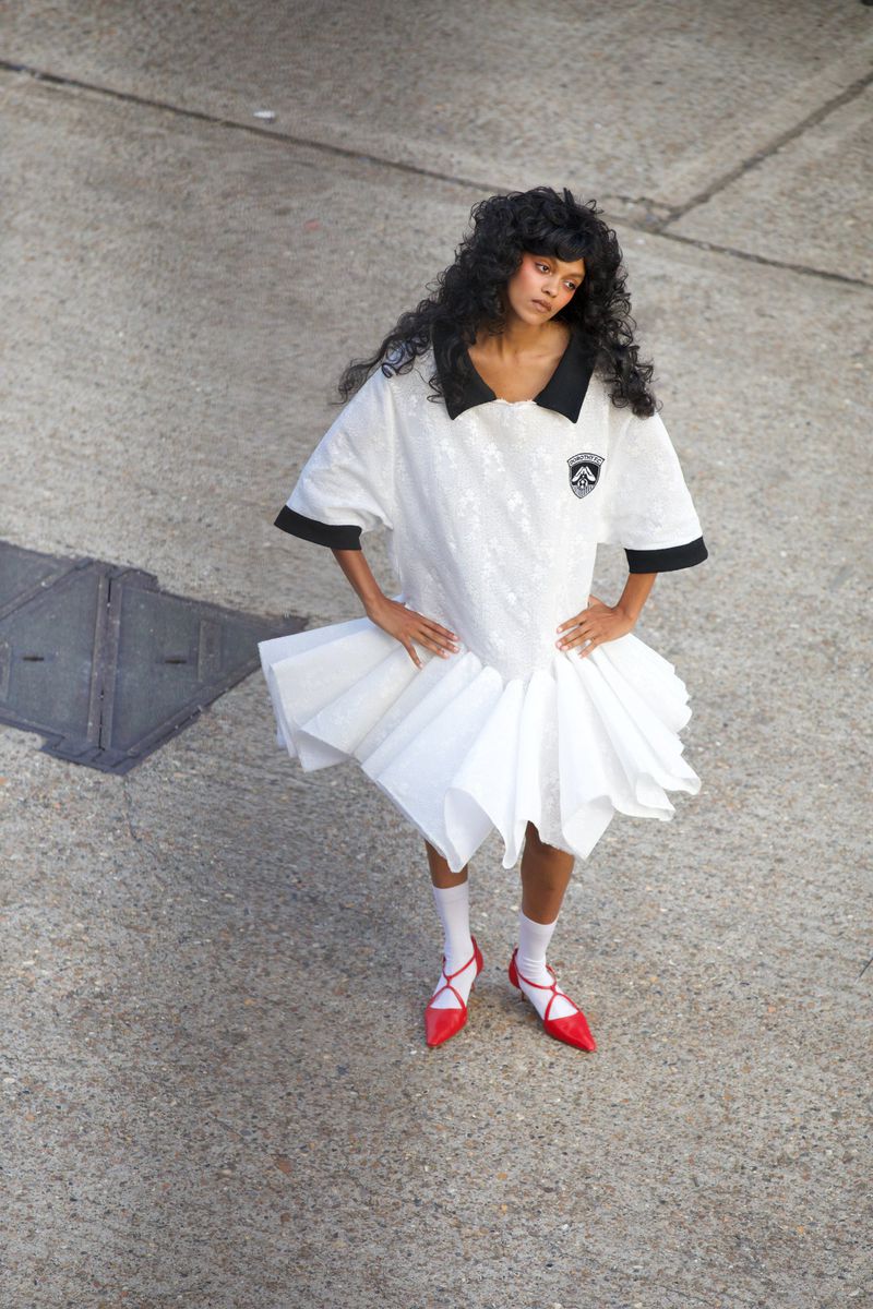 Fashionable woman in a dress that combines a t-shirt top and white layered skirt, posing on concrete with red flats, high-angle.