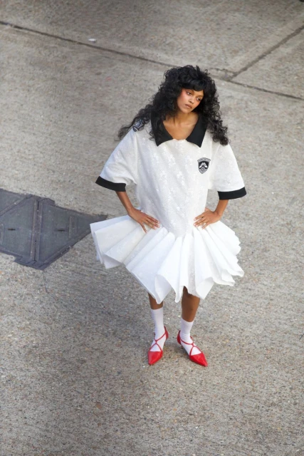 Fashionable woman in a dress that combines a t-shirt top and white layered skirt, posing on concrete with red flats, high-angle.