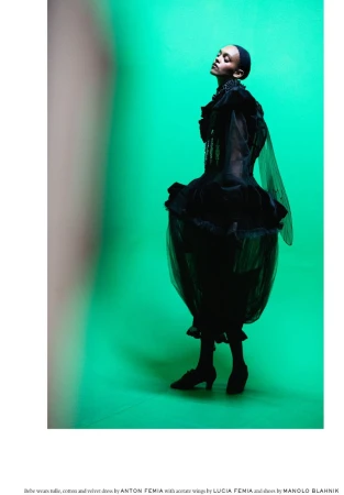 Woman in a black tulle and velvet dress with acetate wings poses against a bright green background in a studio shot for a fashion editorial.