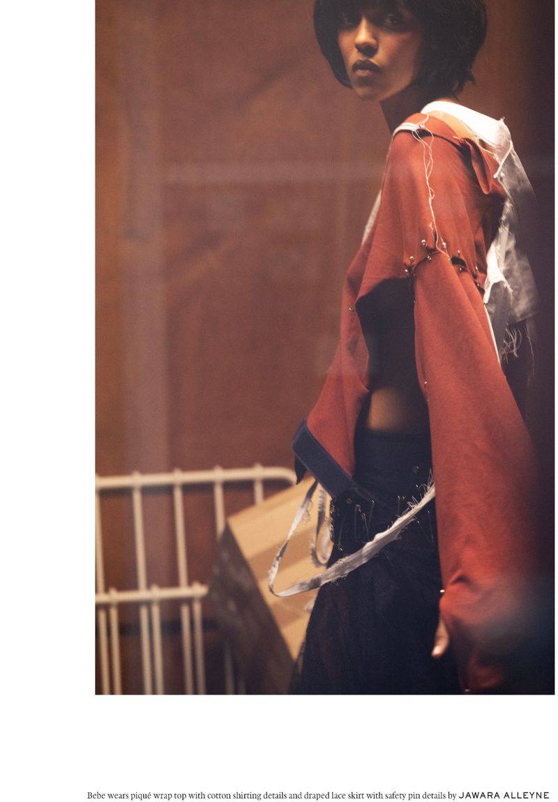 Fashion editorial shot of a woman with short dark hair in an orange wrap top and black lace skirt standing indoors by a railing.