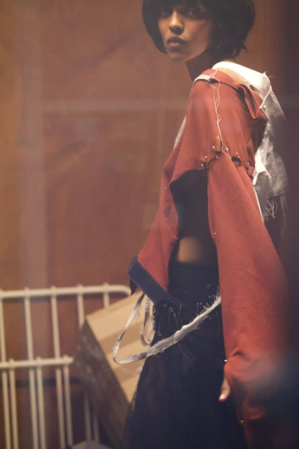 Fashion shot of a female model with short hair wearing a maroon jacket and dark skirt posing indoors in a dimly lit environment.