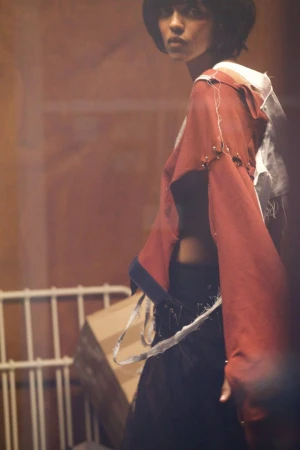 Fashion shot of a female model with short hair wearing a maroon jacket and dark skirt posing indoors in a dimly lit environment.