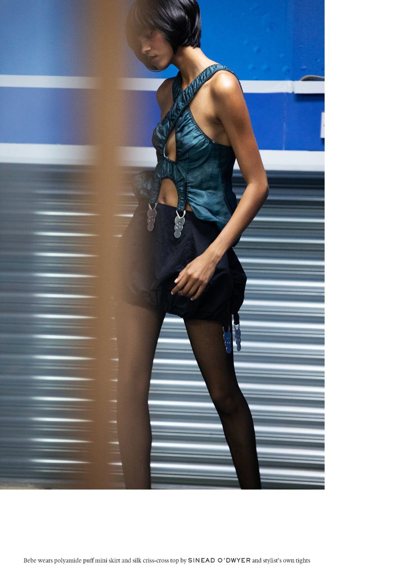 A woman models fashion in an outdoor setting, wearing a silk top and shorts, with a corrugated metal wall as a backdrop, highlighting style.