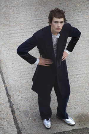 High-angle view of model Noah Bates standing with hands on hips in a long black tailored coat and metallic silver shoes on concrete.