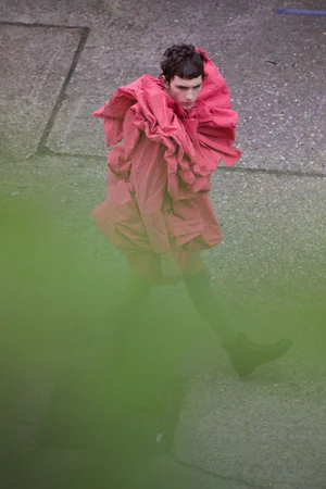 High-angle view of a model walking across textured ground wearing a voluminous ruffled coral red coat with green foreground blur.