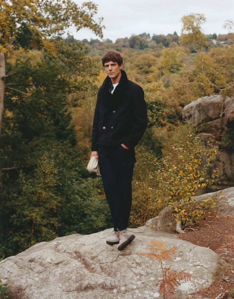 Adult male in a black jacket and trousers stands on a rock outcropping in a wooded area with an autumn-colored forest background.