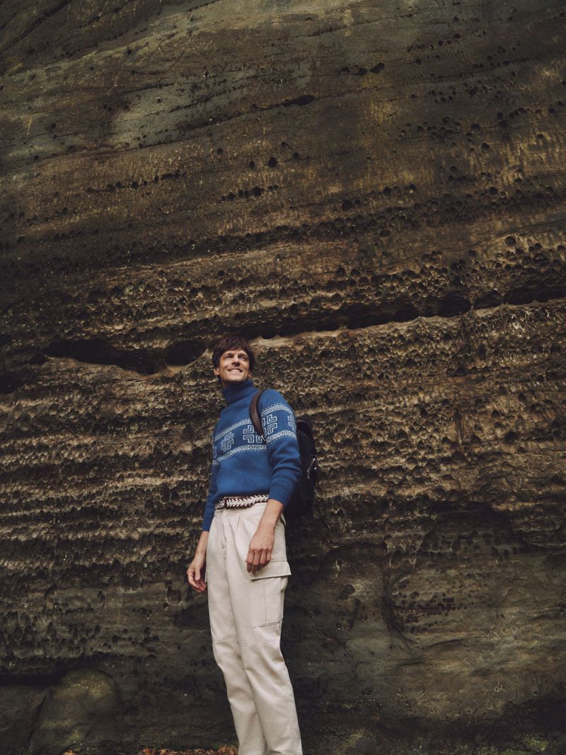 Adult male wearing a blue sweater, backpack and khakis stands outdoors near a weathered rock formation in natural light, looking up.