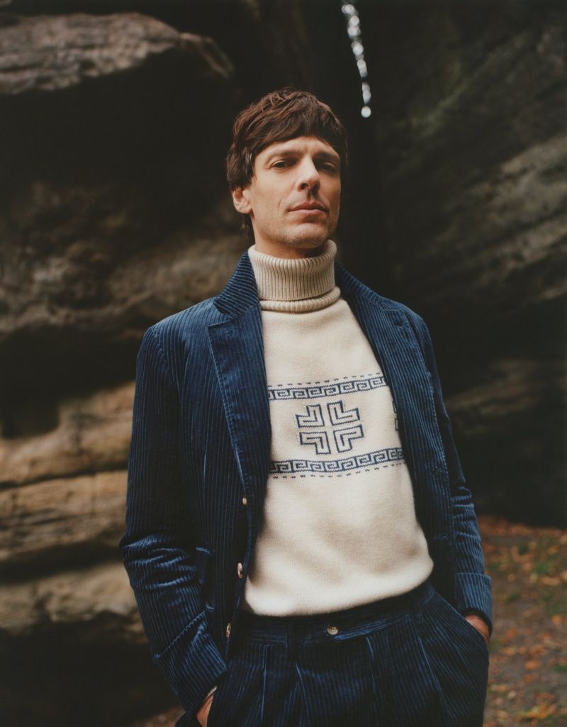 Man in a blue corduroy suit and patterned sweater posing in front of a rock formation in a wooded area, looking at the camera.