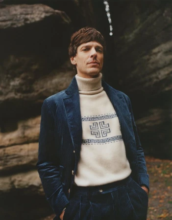 Man in a blue corduroy suit and patterned sweater posing in front of a rock formation in a wooded area, looking at the camera.